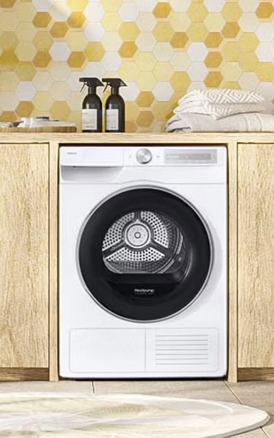 Biała suszarka Samsung w nowoczesnej zabudowie meblowej z jasnego drewna, ustawiona w pralni na tle ściany z żółto-białych heksagonalnych płytek.