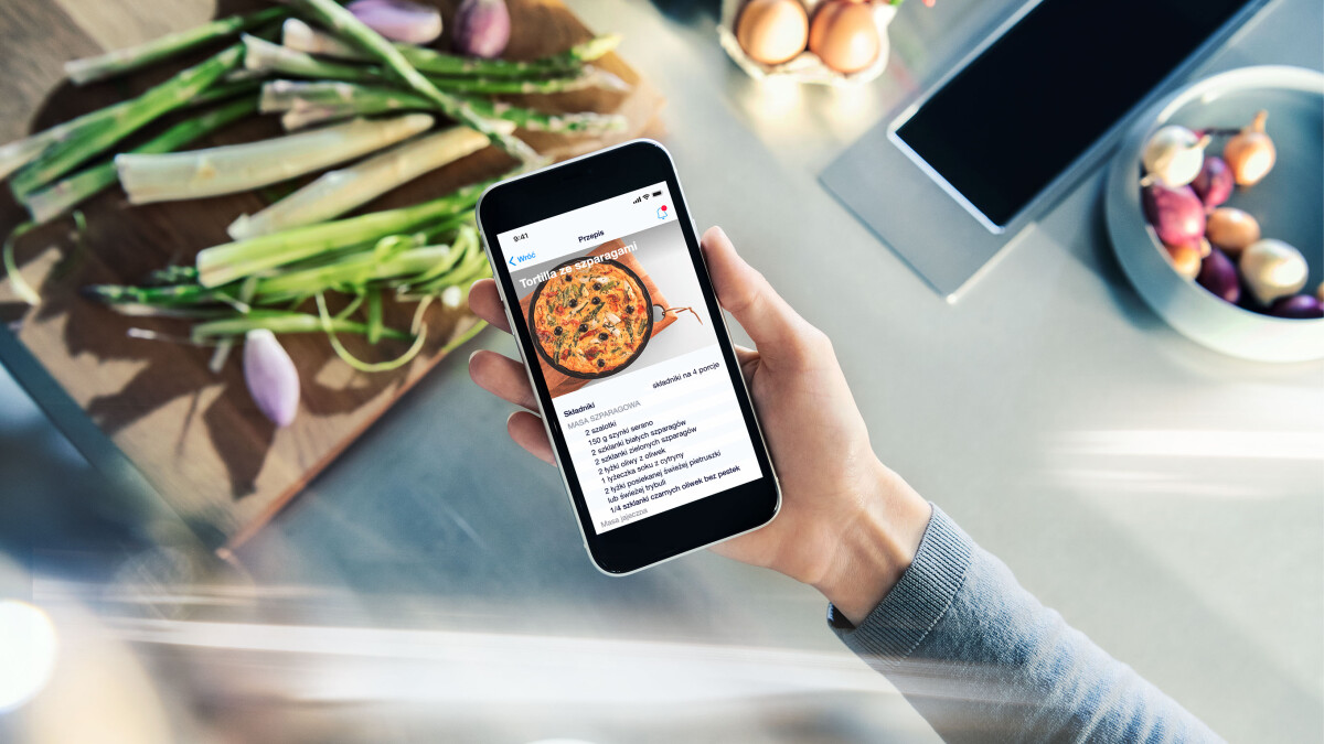 Na pierwszym planie z góry widoczna ręka ze smartfonem i wyświetlaną aplikacją Home Connect. W tle stół kuchenny z produktami.