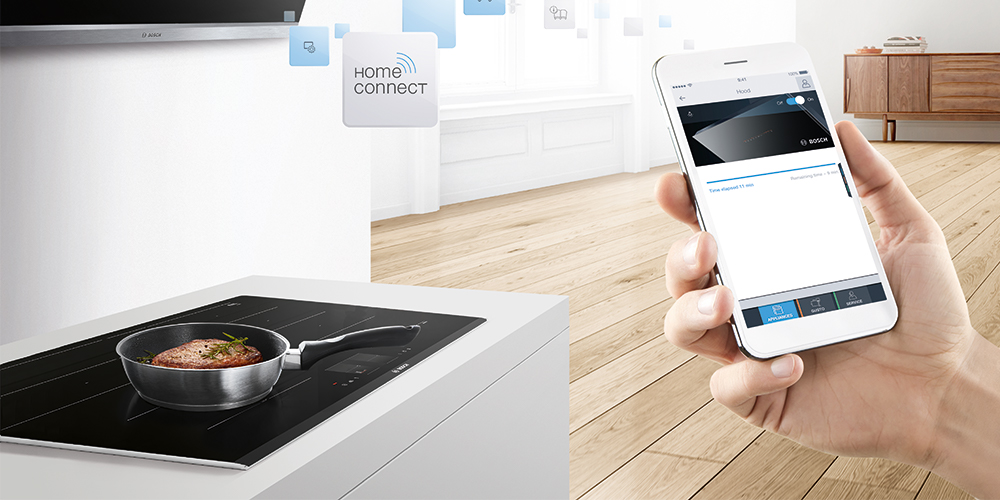 Na pierwszym planie ręka trzymająca smartfona z aplikacją Hom Connect. W oddali okap wiszący nad płytą indukcyjną.